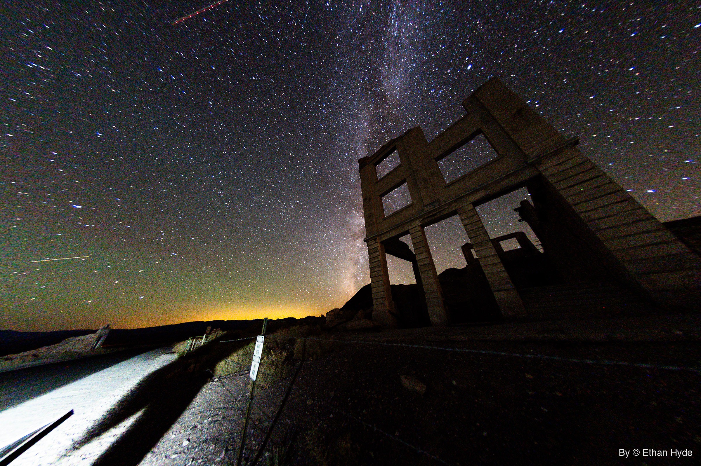 The Milky Way arcs over a ruined concrete bank facade at night, with a red dashed aircraft trail near the top and a lit road at lower left.