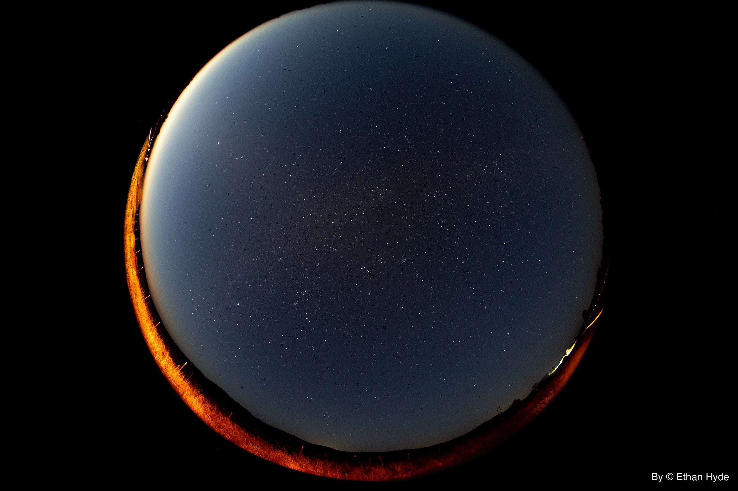 A circular fisheye view of the night sky with a faint band of the Milky Way through the center, ringed by orange-lit grass along the horizon.