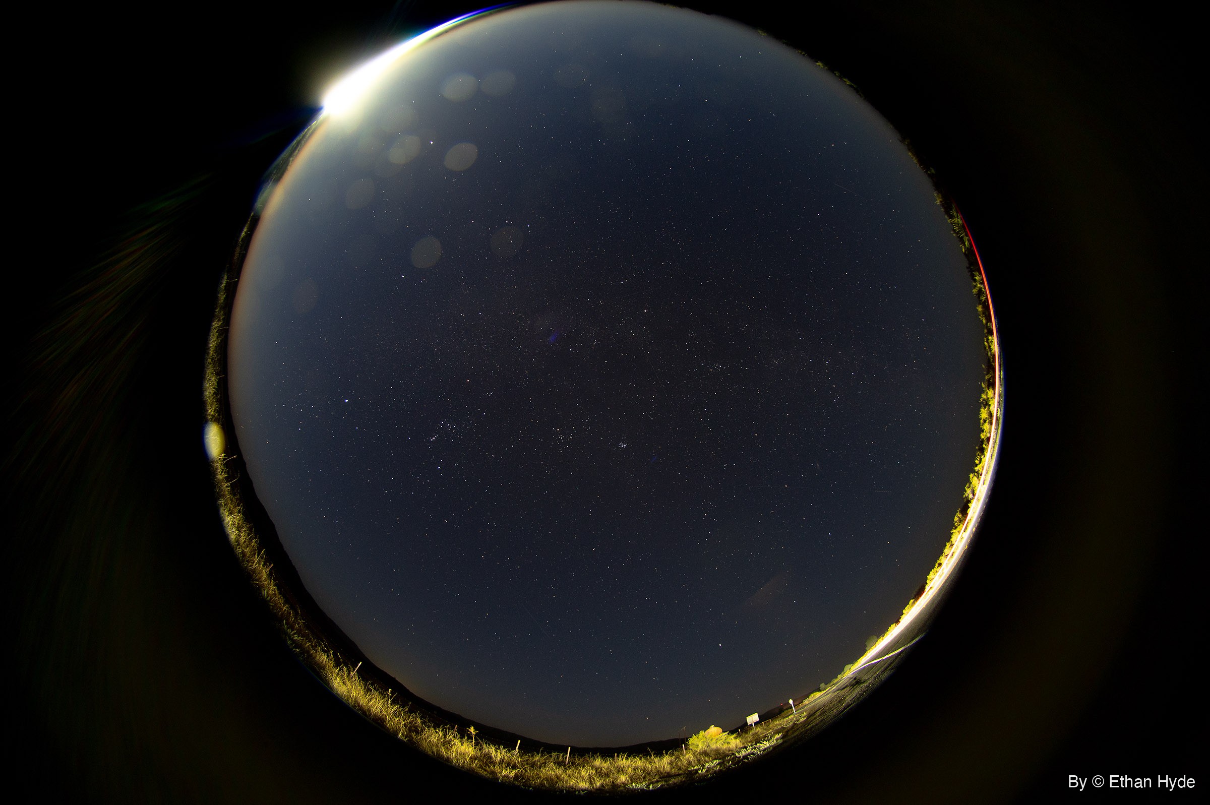 A circular fisheye view of the whole night sky full of faint stars, the moon flaring at the upper-left rim and grass lit gold around the edge.