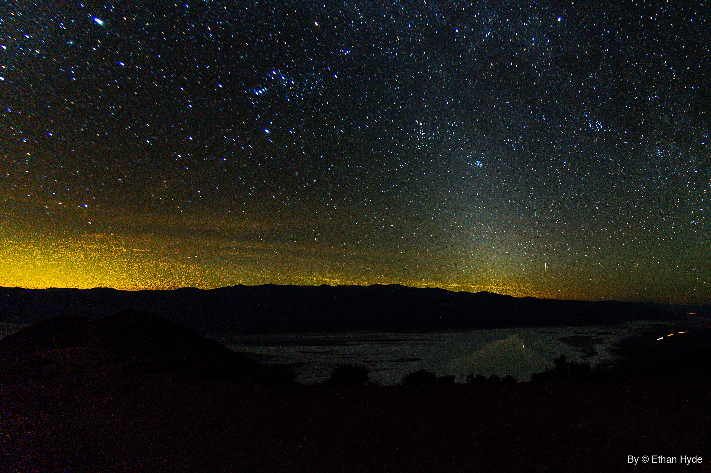 Stars fill the night sky above a dark mountain range, with yellow glow along the left horizon, a thin streak at right, and water on a salt flat.