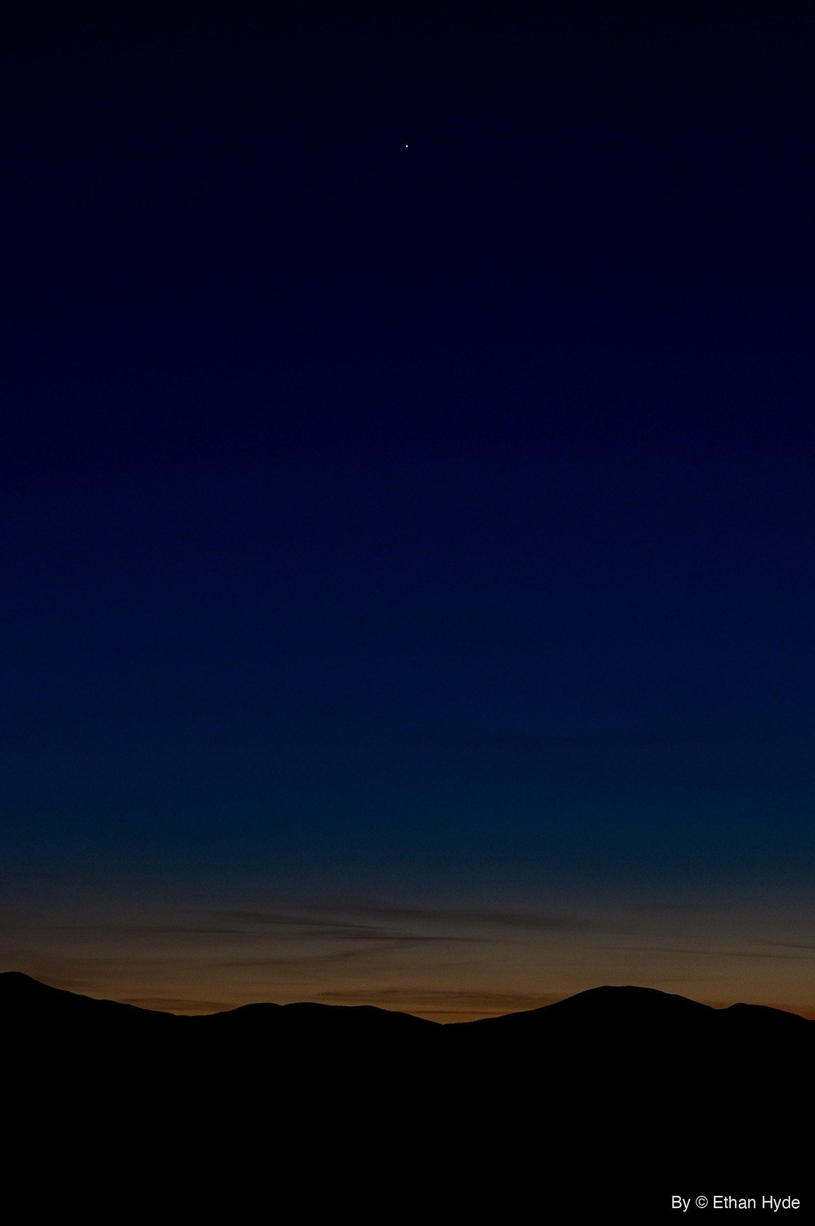 A single bright point of light in a deep blue twilight sky above a black mountain ridgeline, with a thin orange glow along the horizon.