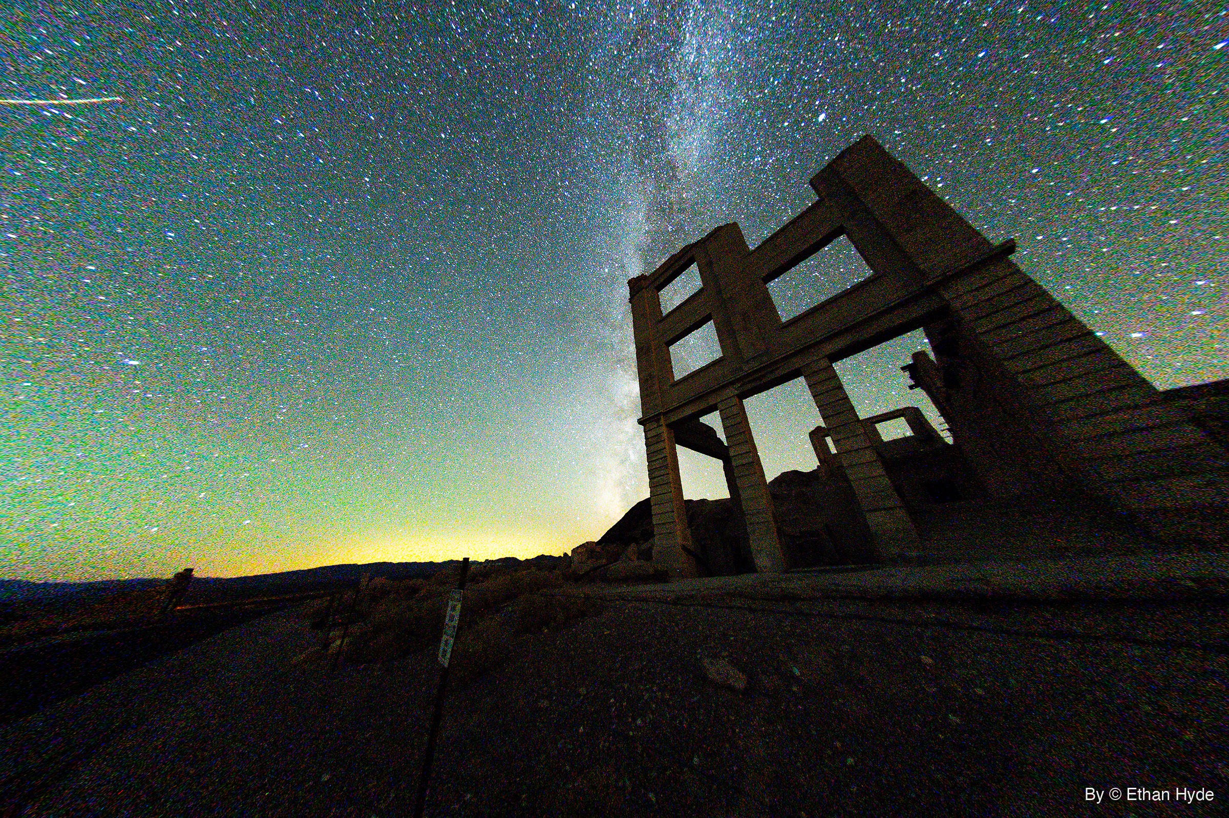 The Milky Way rises behind the roofless concrete facade of a ruined three-story bank building, seen from a low fisheye angle at night.