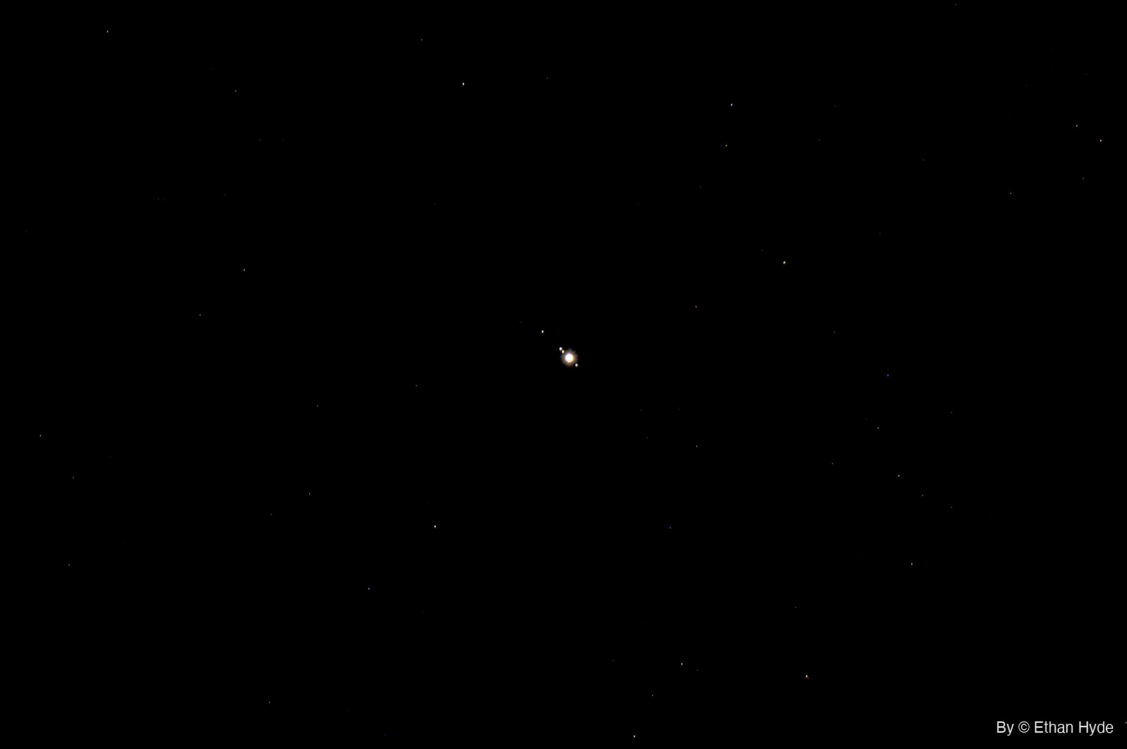 Jupiter shines as a bright point at the center of a black sky, with three tiny moons lined up close beside it and faint stars scattered around.