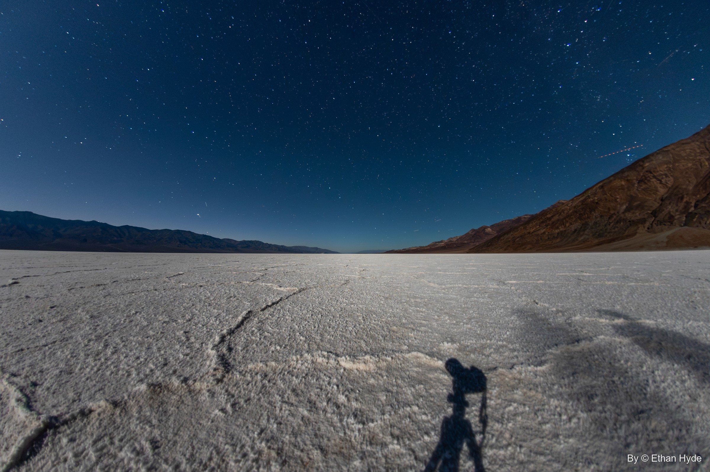 A fisheye night view of stars over the cracked Badwater salt flat, with the photographer's shadow and tripod cast across the crust.