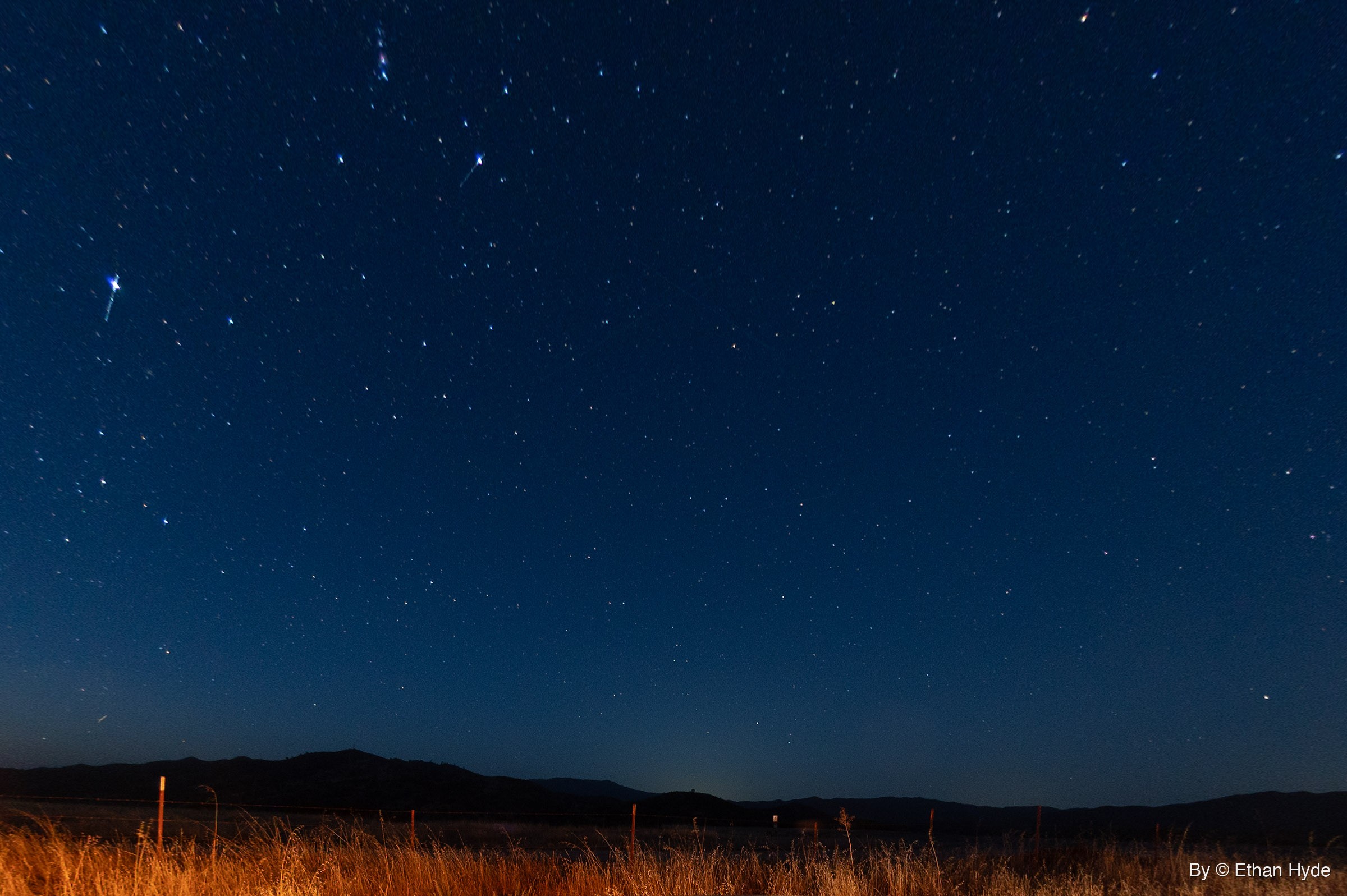A deep blue night sky full of stars above a dark ridgeline, with a strip of lit golden grass and fence posts along the bottom.