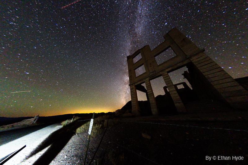 The Milky Way arcs over a ruined concrete bank facade at night, with a red dashed aircraft trail near the top and a lit road at lower left.
