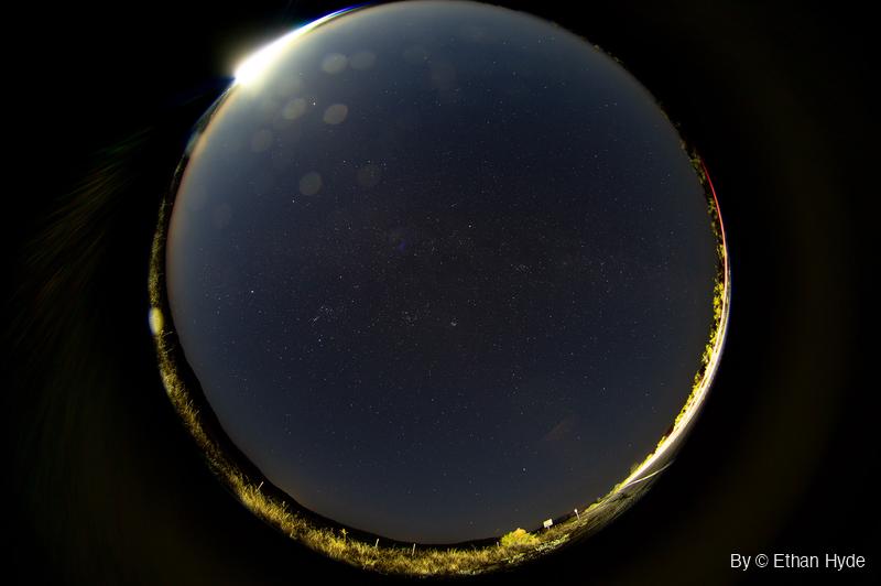 A circular fisheye view of the whole night sky full of faint stars, the moon flaring at the upper-left rim and grass lit gold around the edge.
