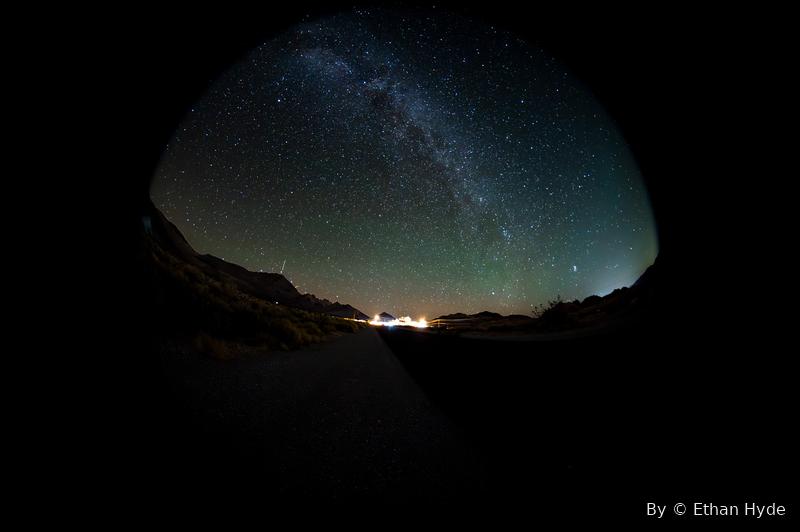 The Milky Way arching over a dark desert canyon, distant town lights on the horizon.