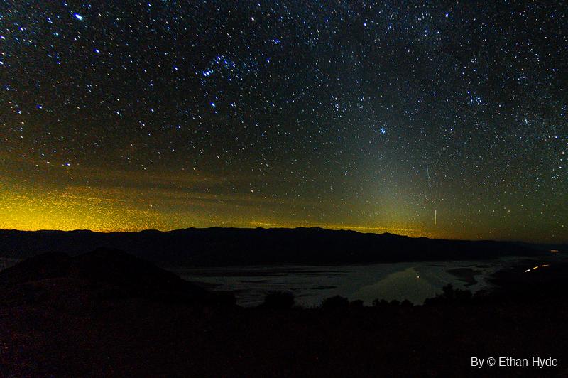 Stars fill the night sky above a dark mountain range, with yellow glow along the left horizon, a thin streak at right, and water on a salt flat.