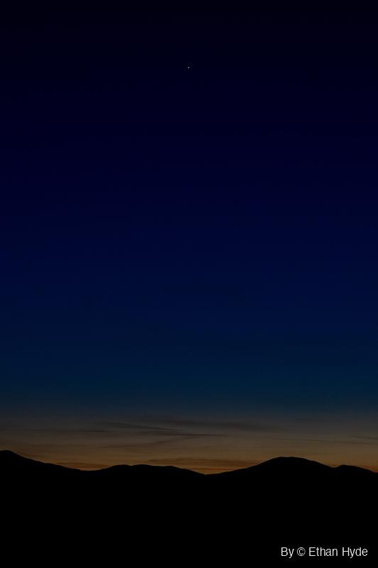 A single bright point of light in a deep blue twilight sky above a black mountain ridgeline, with a thin orange glow along the horizon.