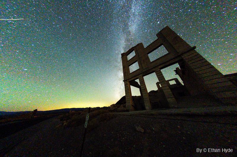 The Milky Way rises behind the roofless concrete facade of a ruined three-story bank building, seen from a low fisheye angle at night.
