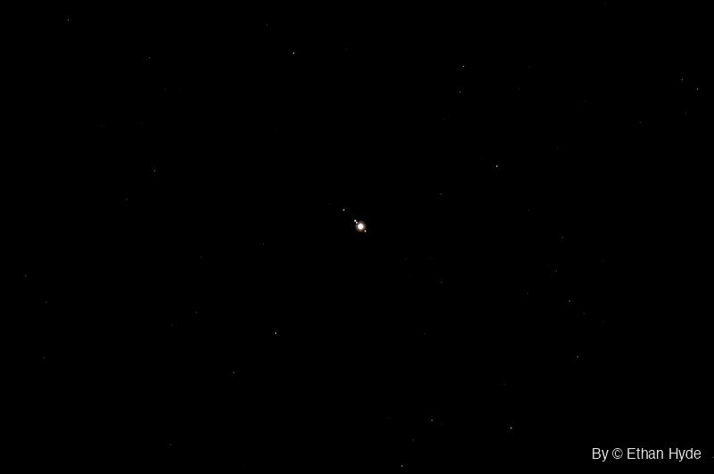 Jupiter shines as a bright point at the center of a black sky, with three tiny moons lined up close beside it and faint stars scattered around.