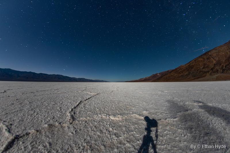 A fisheye night view of stars over the cracked Badwater salt flat, with the photographer's shadow and tripod cast across the crust.