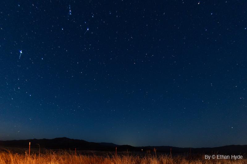 A deep blue night sky full of stars above a dark ridgeline, with a strip of lit golden grass and fence posts along the bottom.