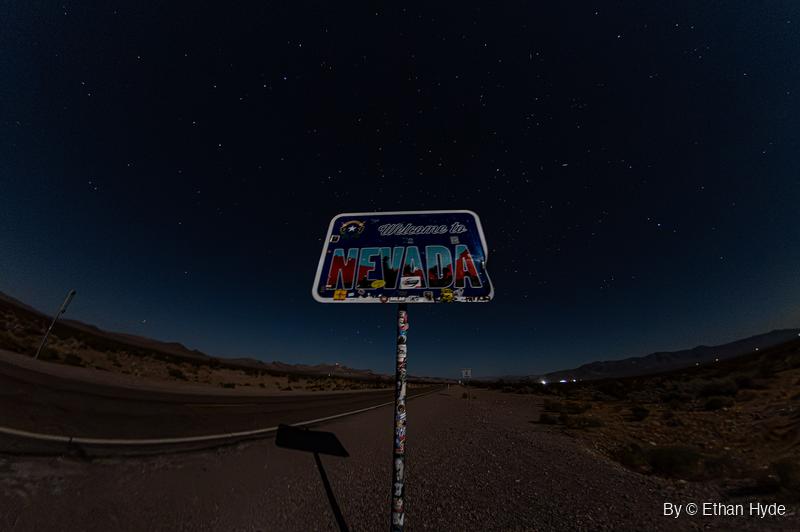 A sticker-covered Welcome to Nevada sign stands beside an empty desert highway at night under a star-filled sky.