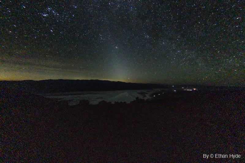 A star-filled night sky over a dark mountain ridge, with a faint cone of light rising above a pale salt flat holding water.