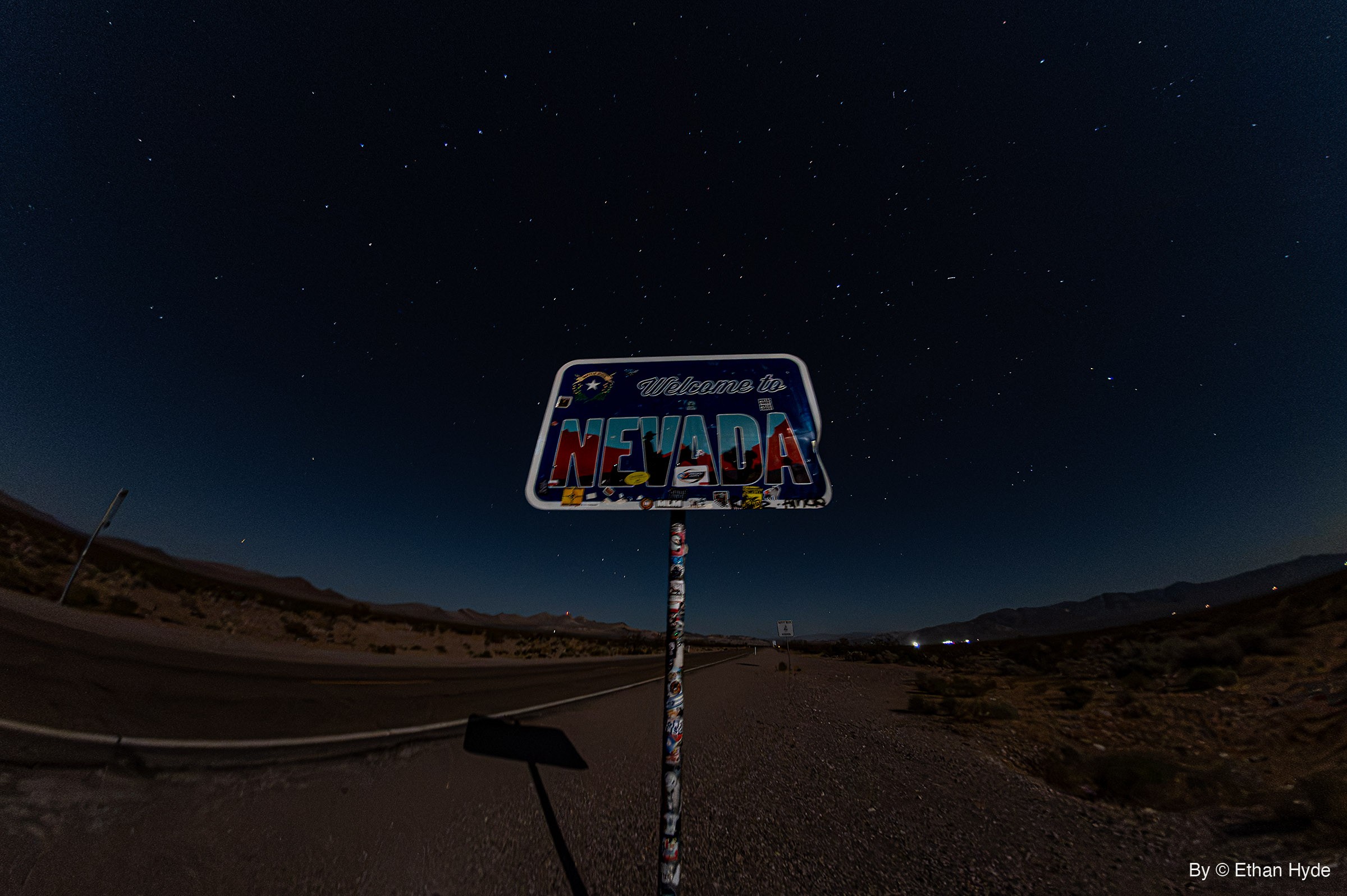A sticker-covered Welcome to Nevada sign stands beside an empty desert highway at night under a star-filled sky.
