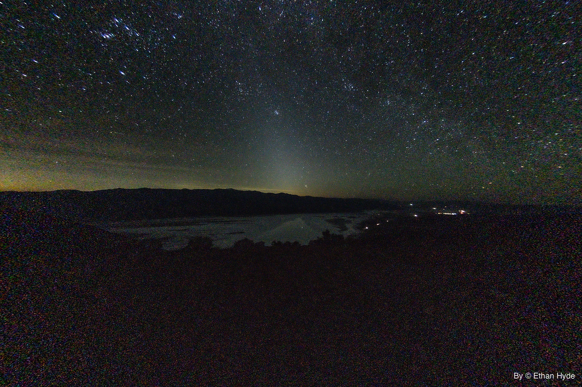A star-filled night sky over a dark mountain ridge, with a faint cone of light rising above a pale salt flat holding water.