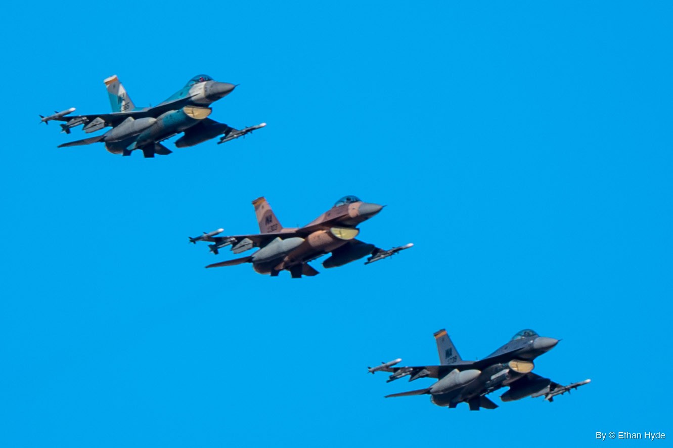 Three F-16 fighters in blue-gray, tan-brown and dark gray camouflage fly in a diagonal line against a clear blue sky.