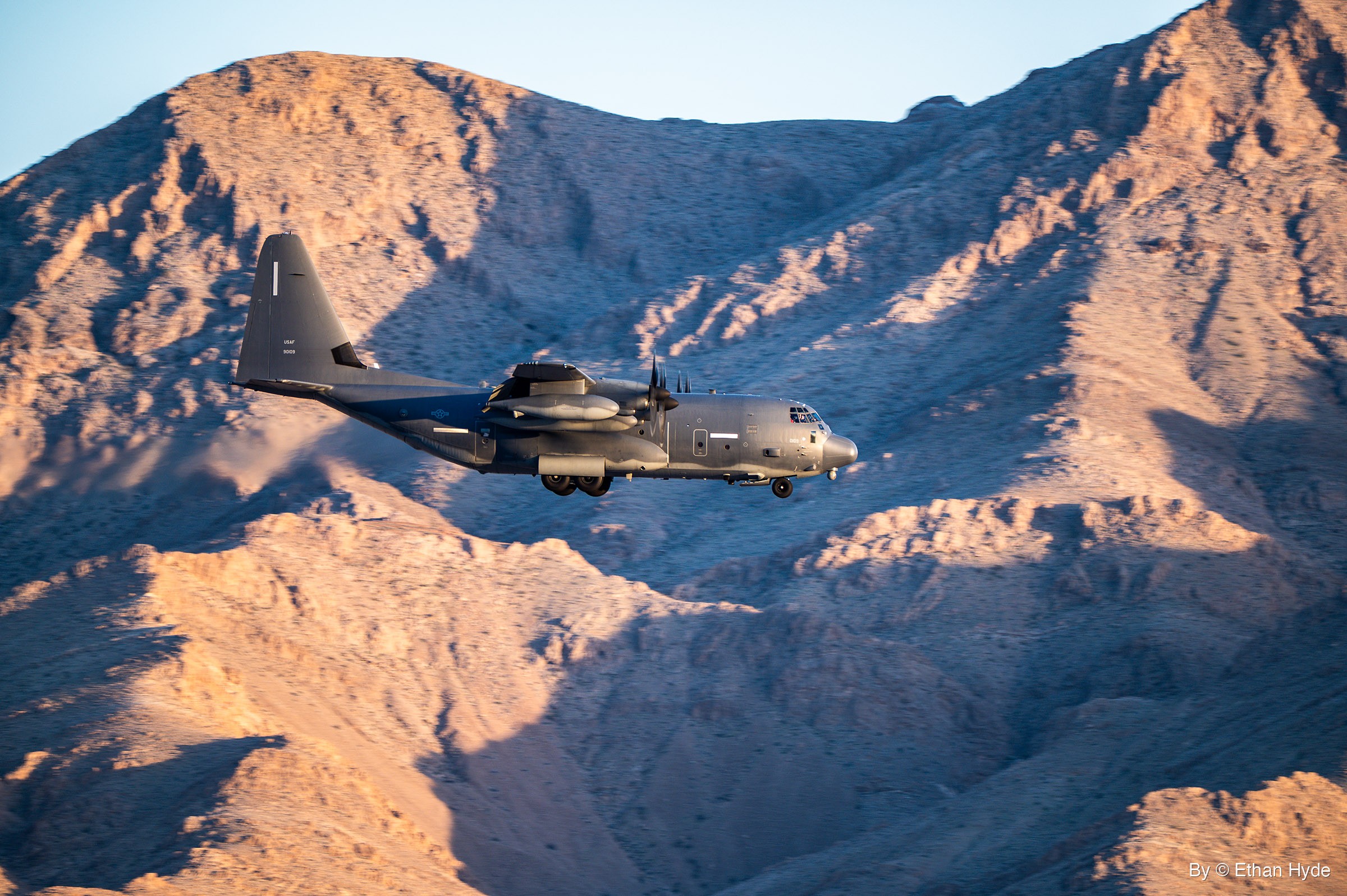 A dark gray four-engine turboprop military transport flies with its landing gear down in front of sunlit, eroded desert mountains.
