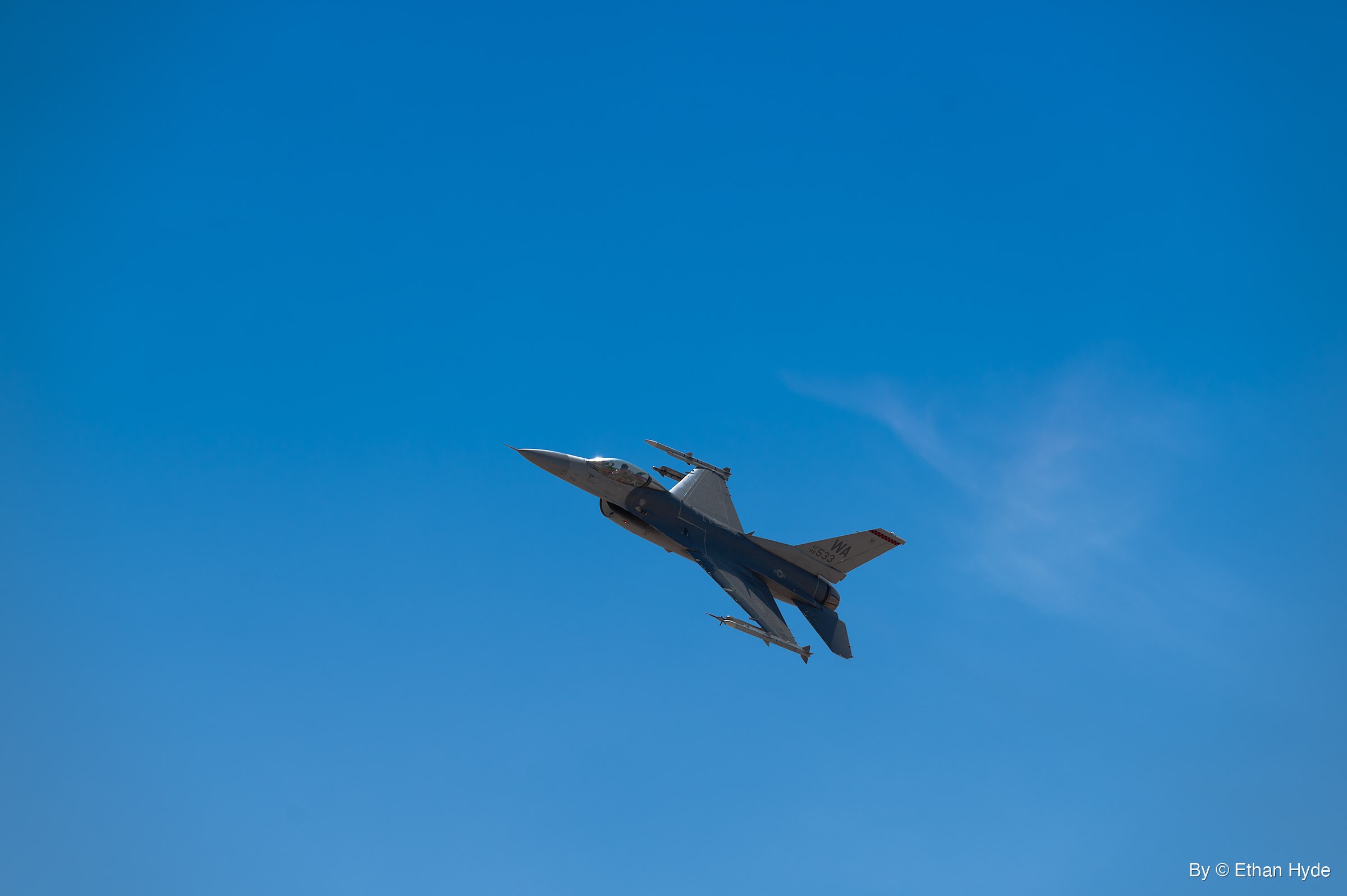 A gray F-16 fighter jet banks against a clear blue sky, a missile on each wingtip, with a thin streak of cloud to the right.