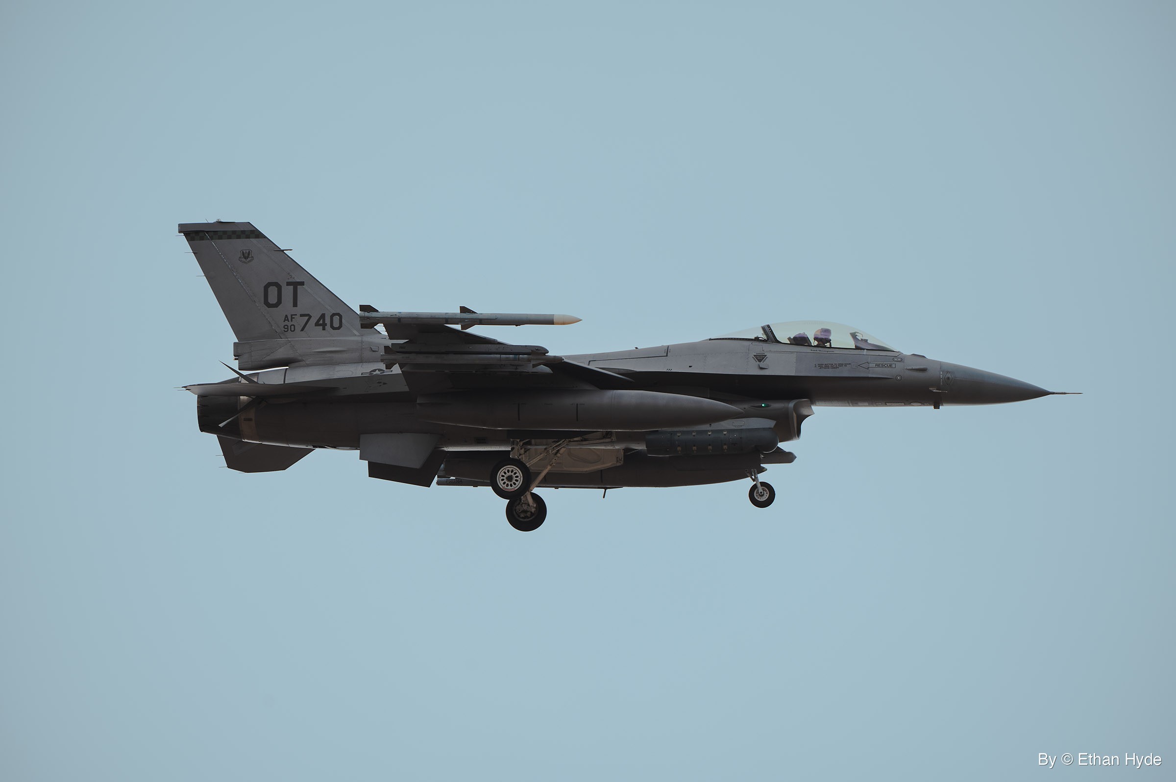 A gray F-16 fighter jet flies level in side profile with its landing gear down against a pale blue sky, pilot visible in the canopy.