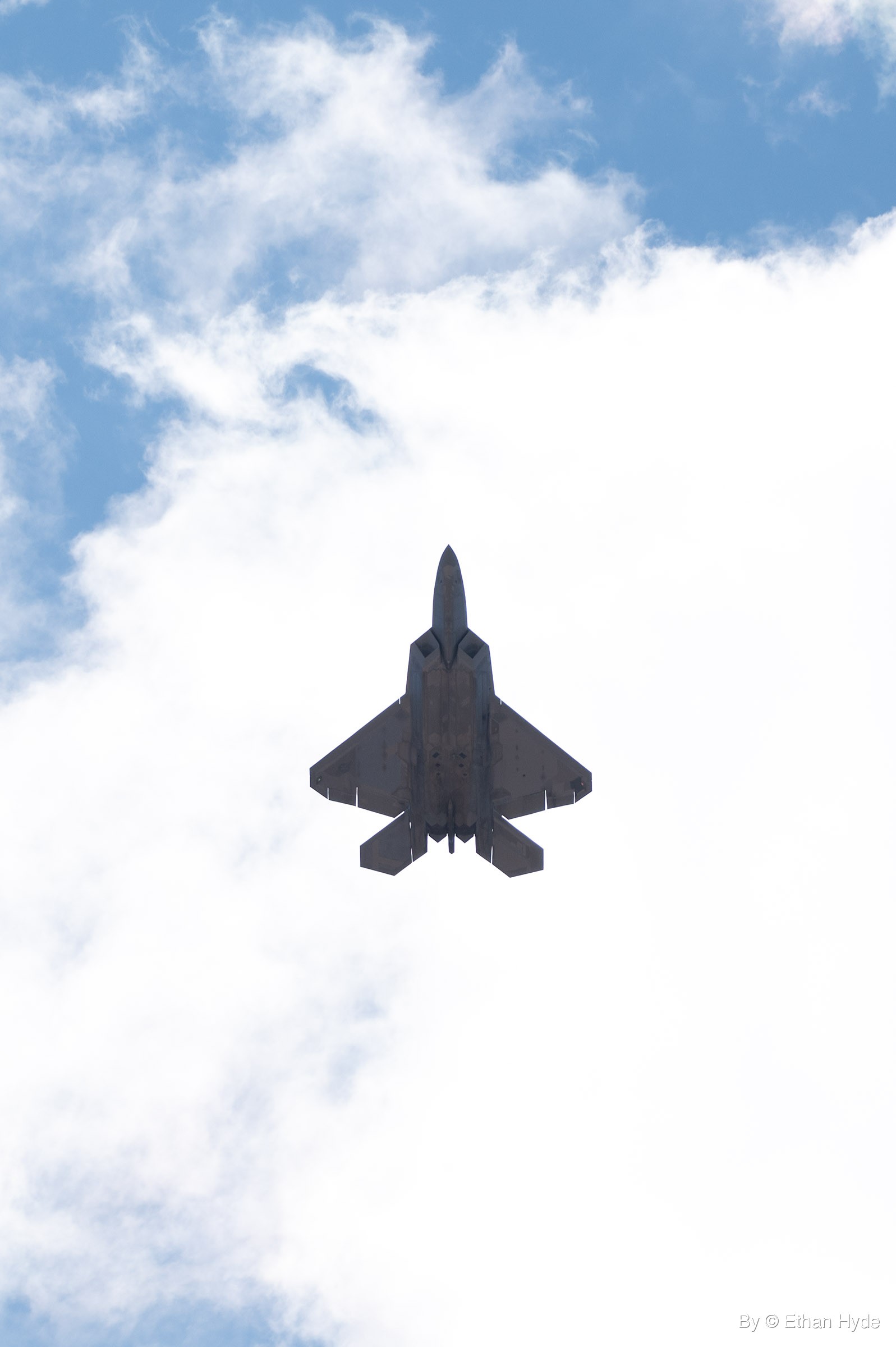 An F-22 fighter jet seen from directly below, crossing a bank of bright white cloud with patches of blue sky above.
