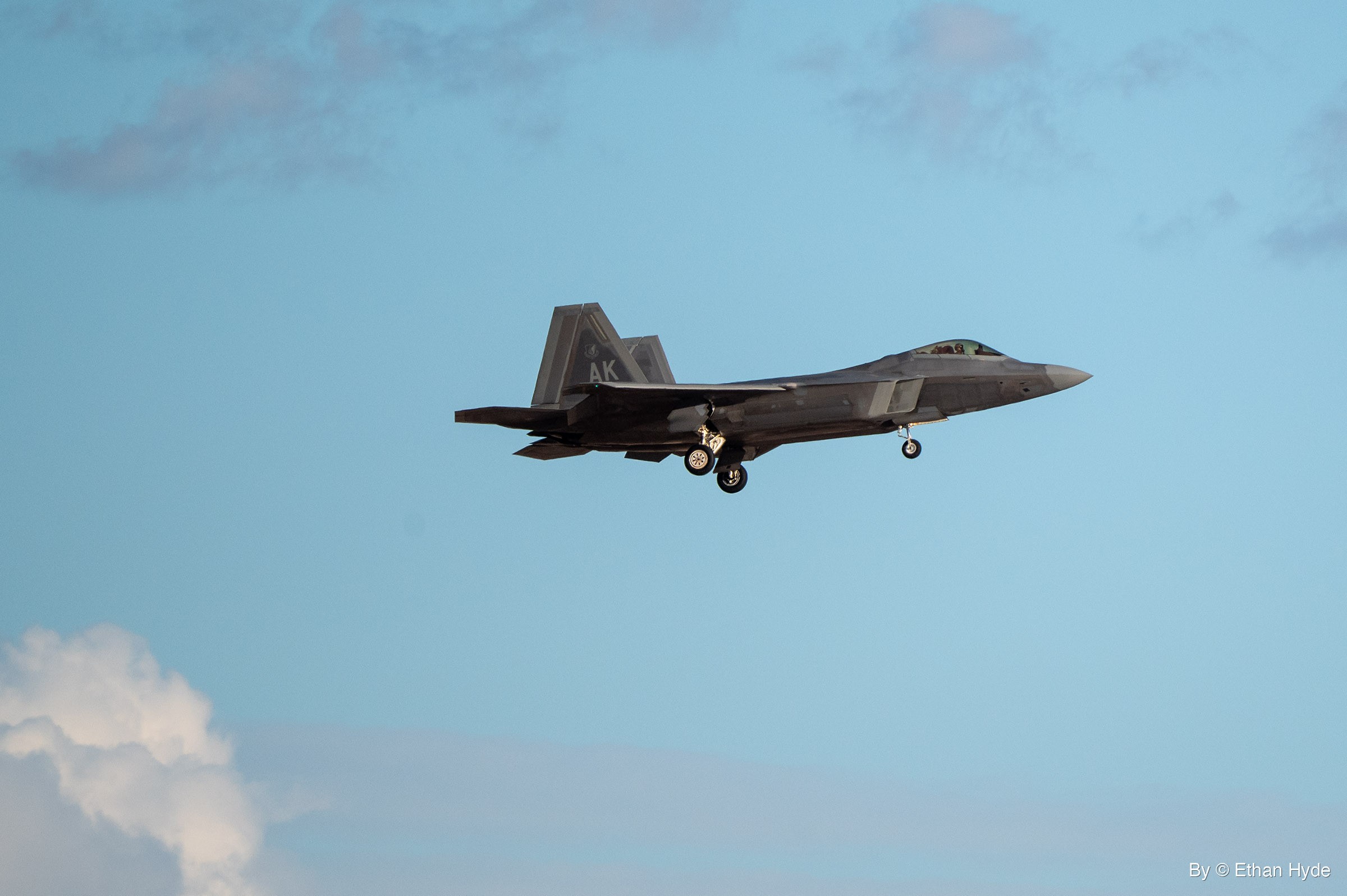 An F-22 Raptor flies on approach with its landing gear down against a pale blue sky, a white cloud at lower left.
