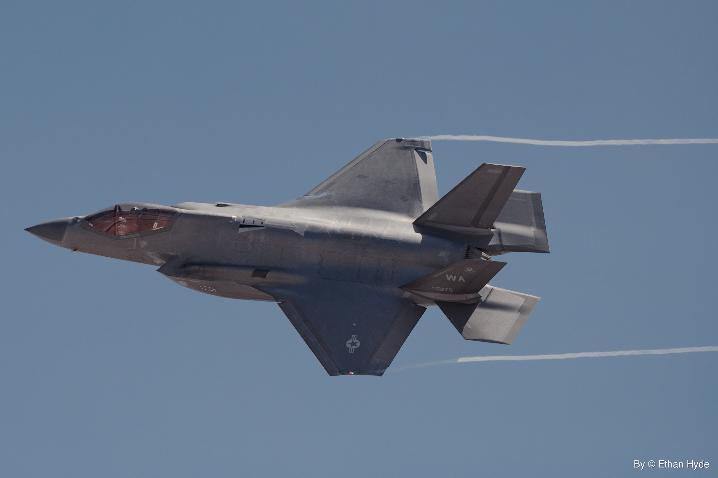 A gray F-35 fighter jet flies right to left in a banked turn with its underside showing, thin white vapor trails streaming behind it.