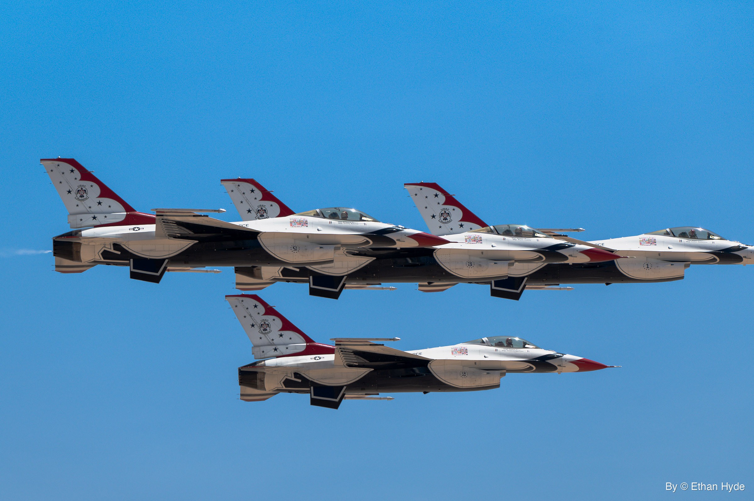 Four F-16 fighter jets in red, white and blue demonstration paint fly in tight formation, three overlapping in a line and one below, against blue sky.