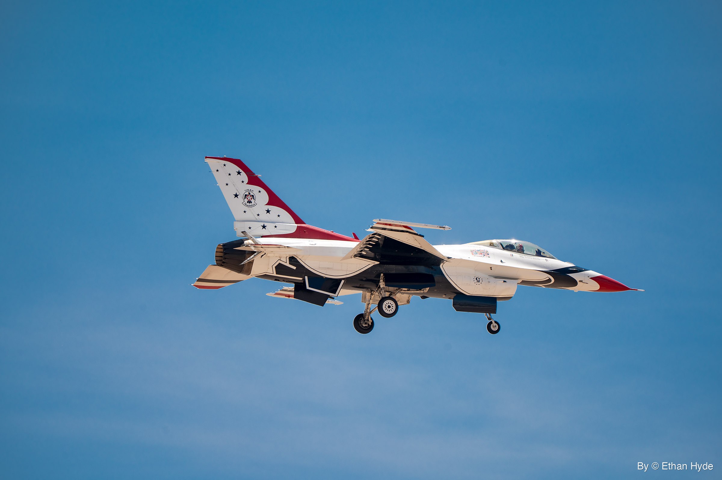 A single F-16 in red, white and blue demonstration paint flies with its landing gear extended against a hazy blue sky.