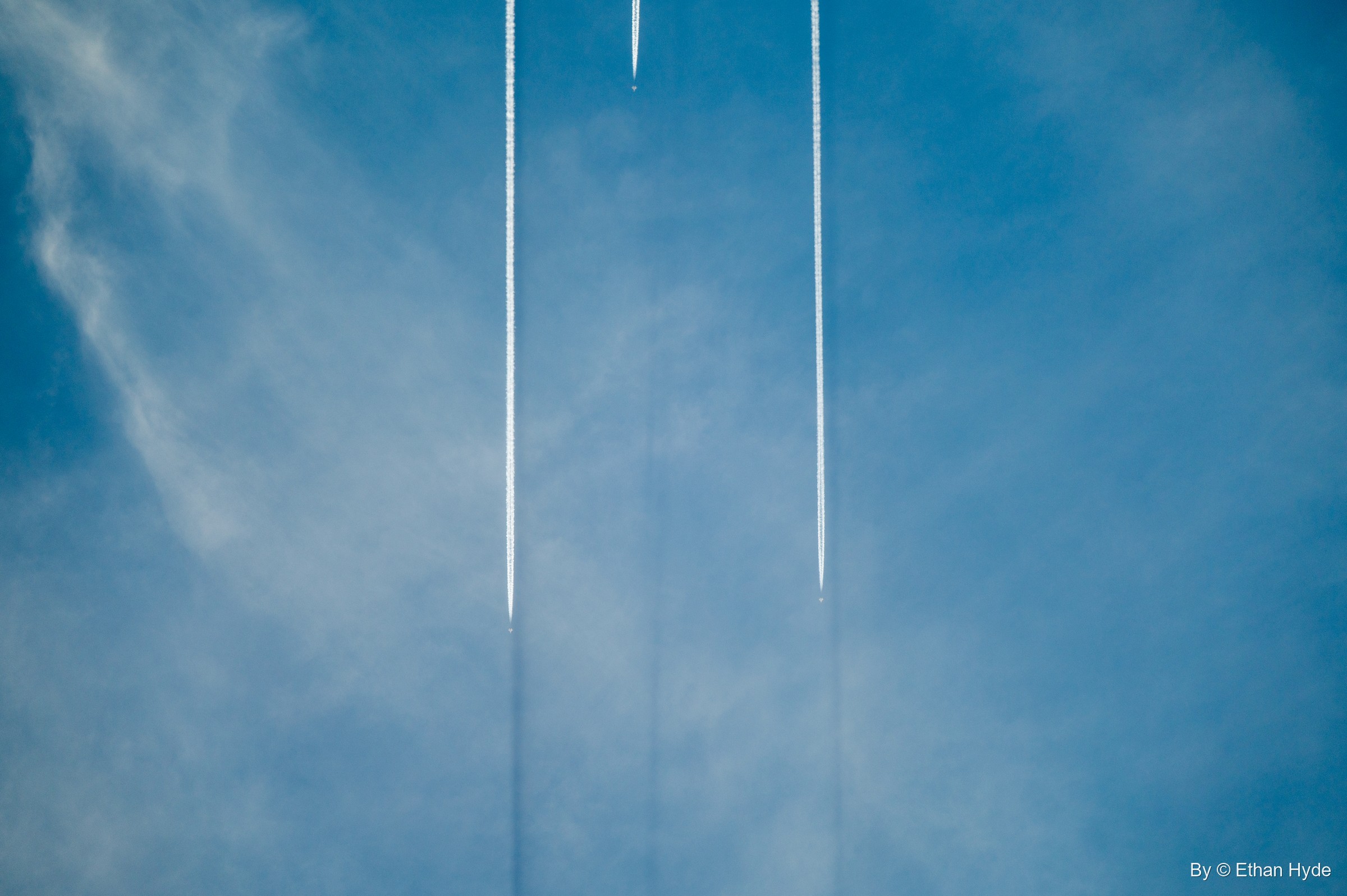 Two straight white contrails run side by side up a blue sky streaked with thin cloud.