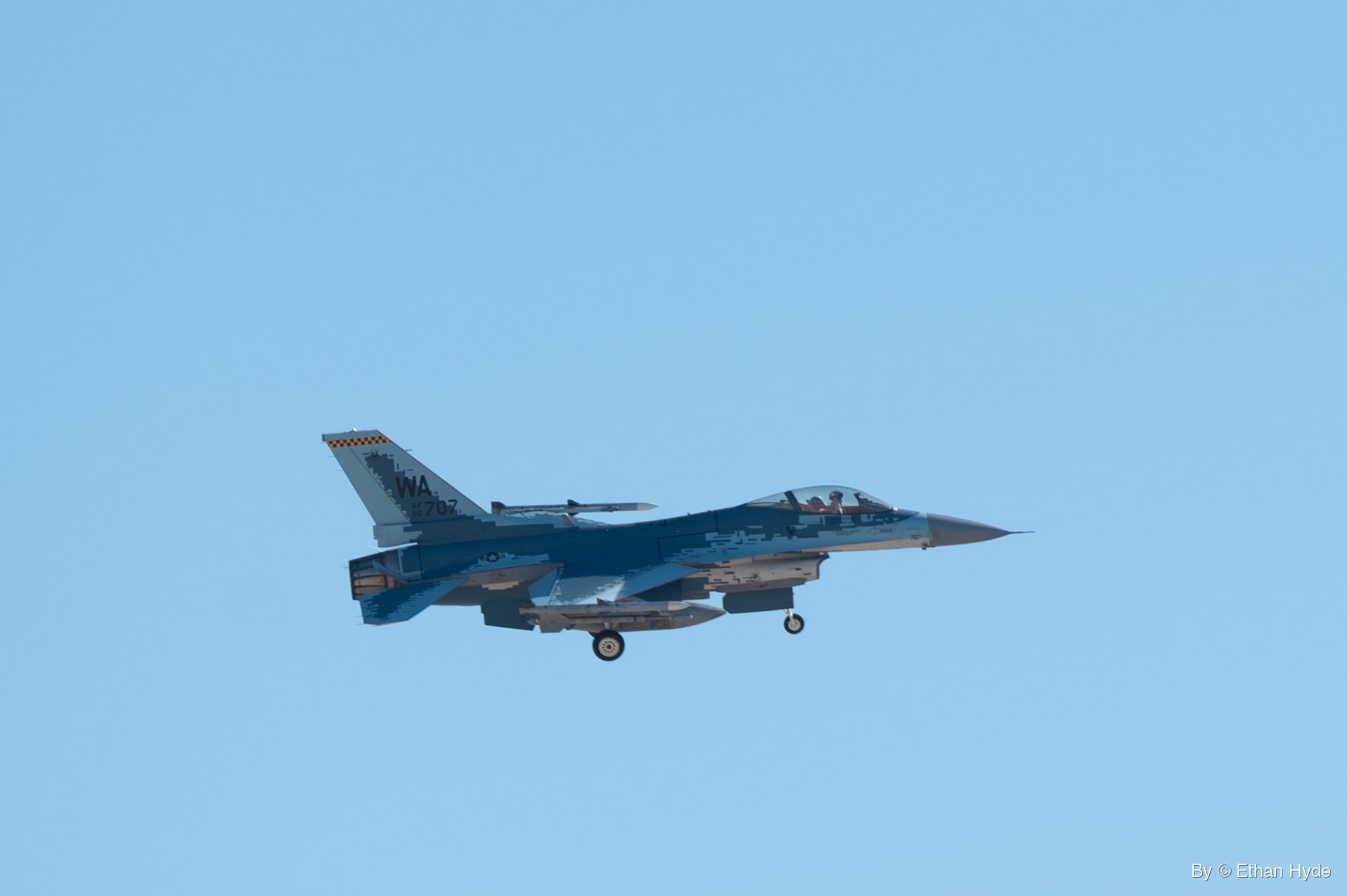 An F-16 in blue and gray pixelated camouflage flies with its landing gear down against a clear blue sky.