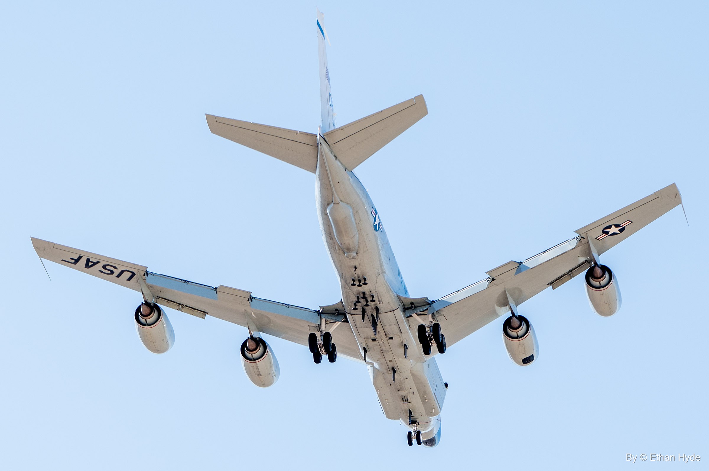 A white four-engine US Air Force RC-135 passes directly overhead with landing gear extended, USAF lettering and star insignia under the wings.