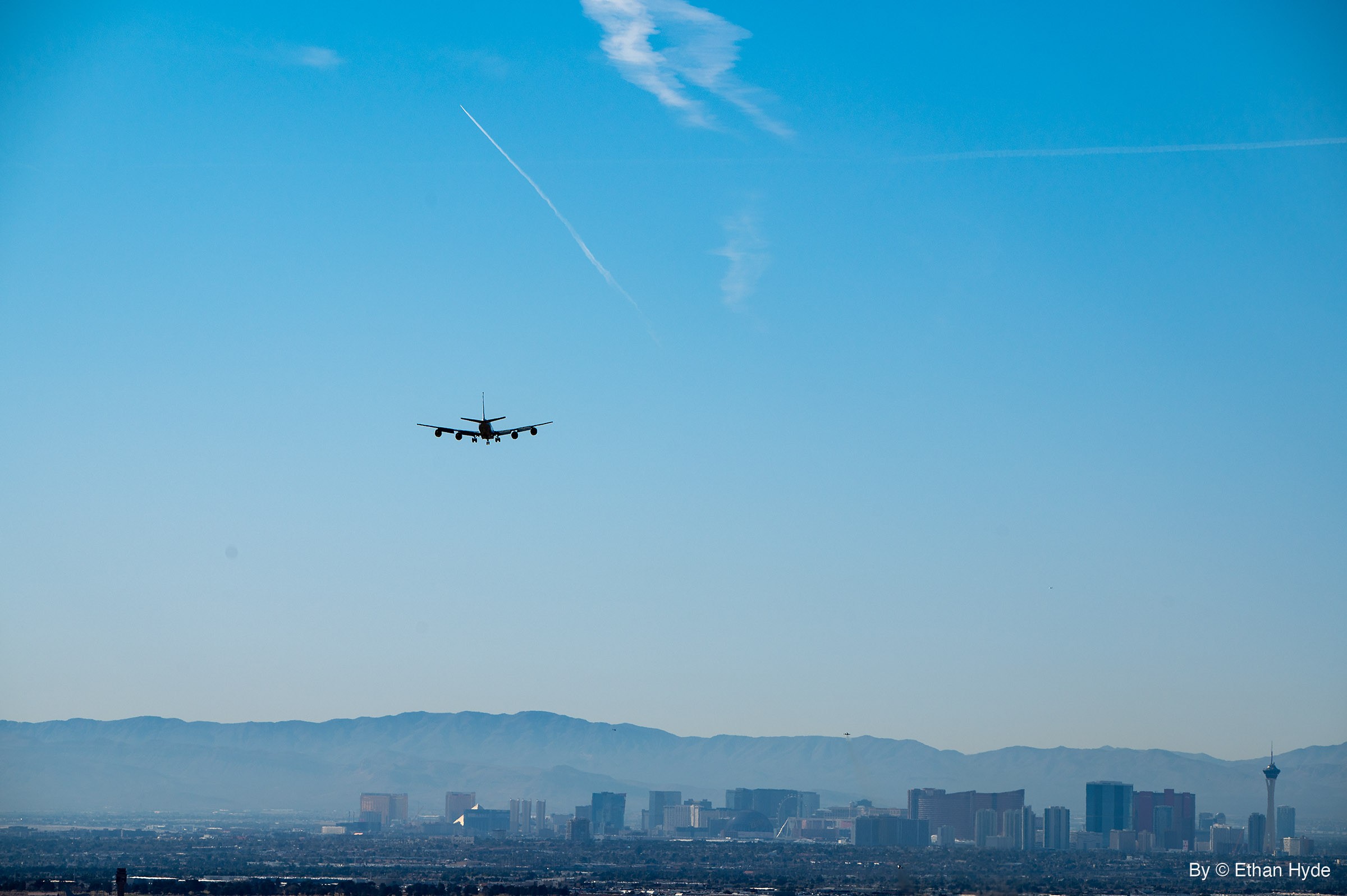 A four-engine RC-135 with landing gear down flies above a hazy skyline of high-rise towers, with a mountain range behind.