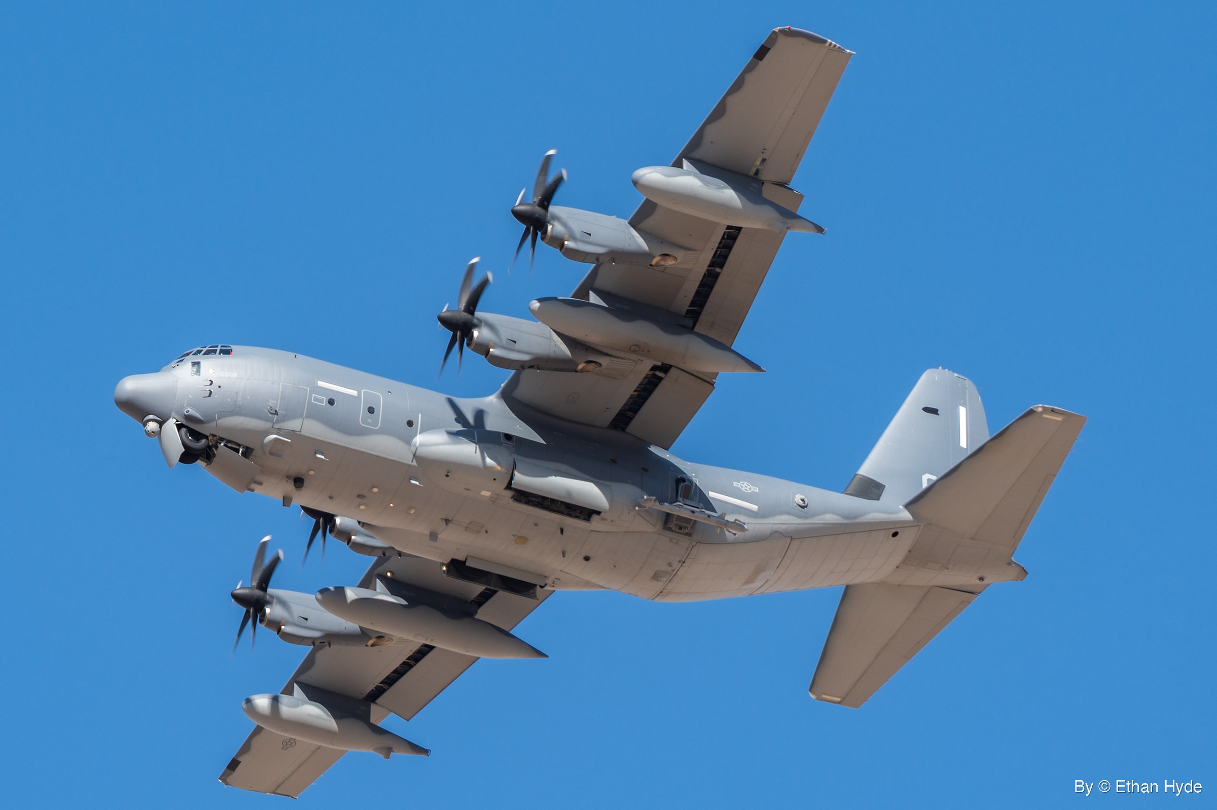 A gray four-engine military turboprop with six-bladed propellers, underwing fuel tanks and outer-wing pods passes overhead against clear blue sky.