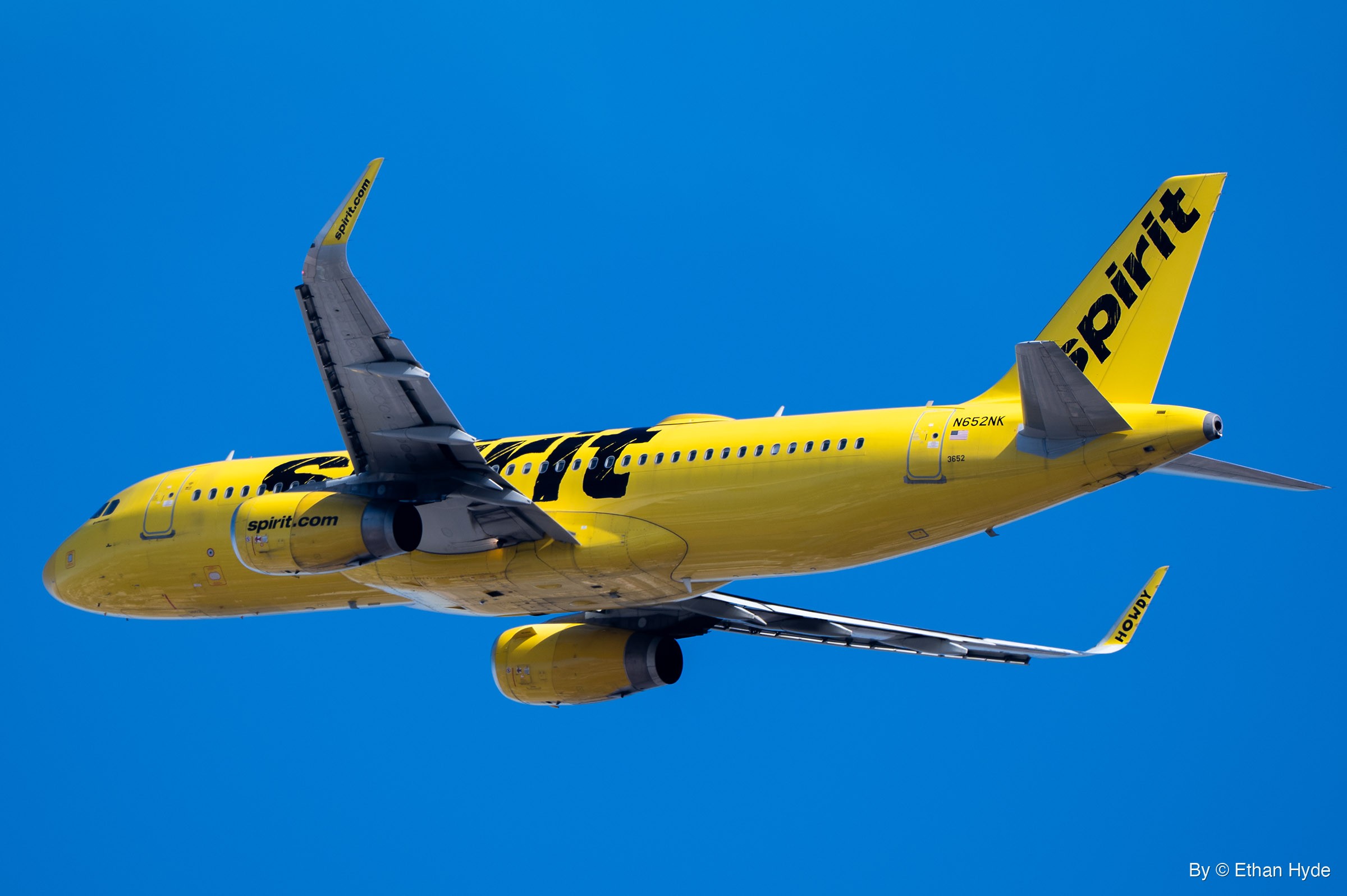 A yellow Spirit Airlines Airbus A320 rolls into a steep bank, seen from below with one wing raised against deep blue sky.
