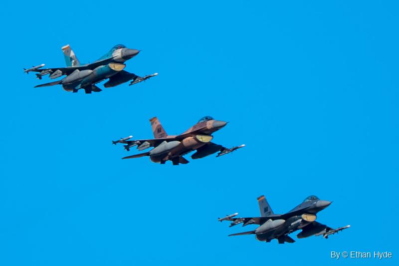 Three F-16 fighters in blue-gray, tan-brown and dark gray camouflage fly in a diagonal line against a clear blue sky.