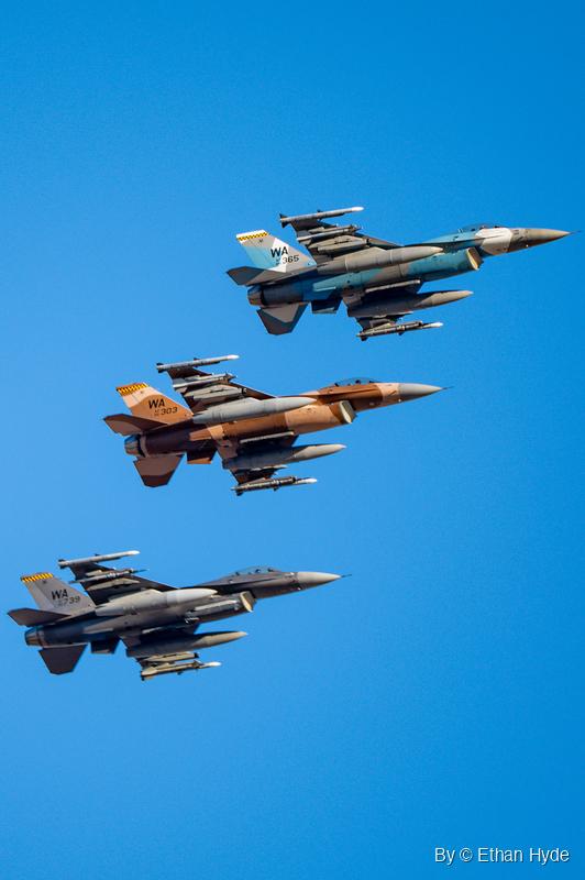 Three camouflaged F-16 fighters in blue-gray, tan-brown and dark gray fly past overhead in a stepped line against blue sky.