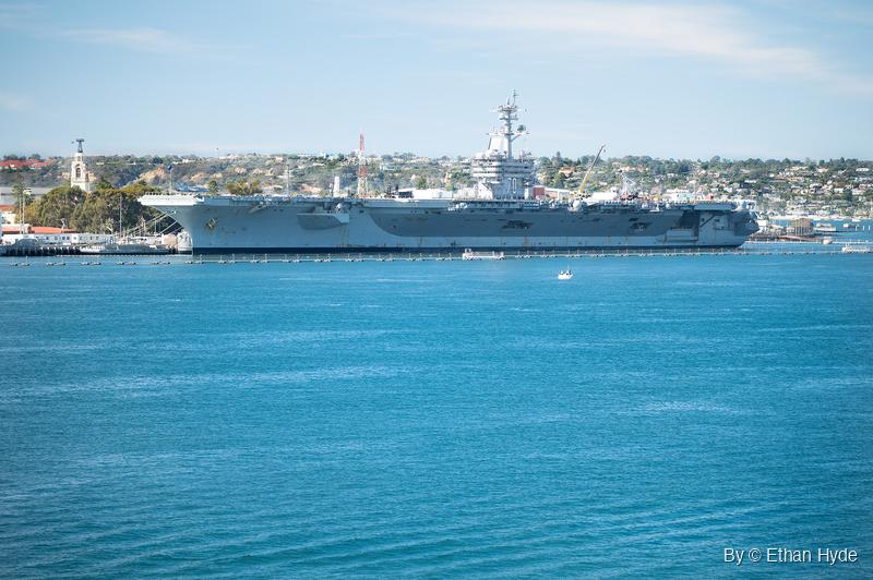 Gray aircraft carrier moored at a pier behind a line of floating barriers, with a small boat on blue water and hillside homes behind.