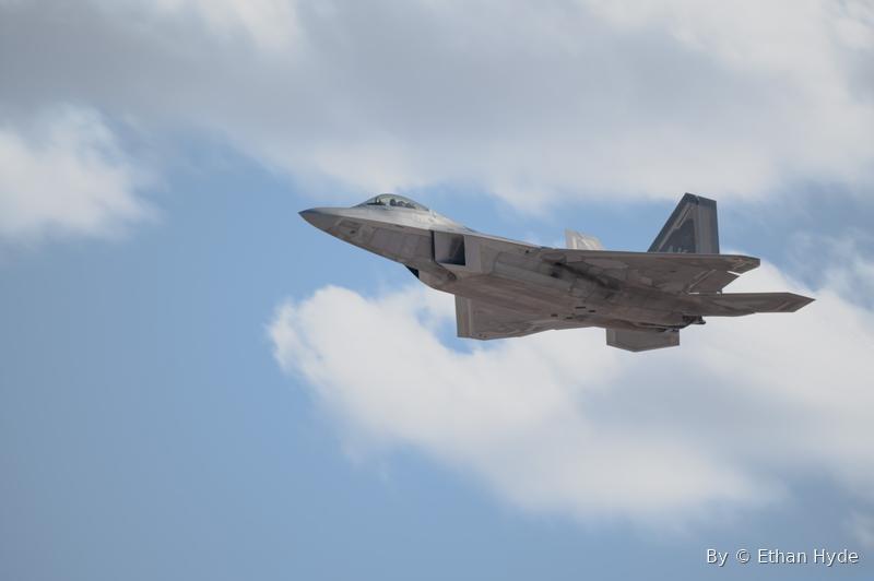 A fighter jet climbing past scattered white clouds in a blue sky.