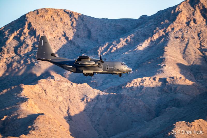 A dark gray four-engine turboprop military transport flies with its landing gear down in front of sunlit, eroded desert mountains.