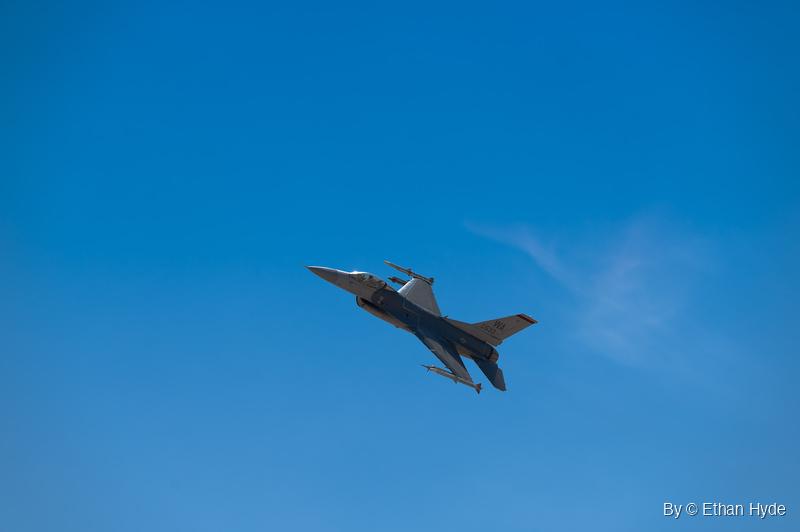 A gray F-16 fighter jet banks against a clear blue sky, a missile on each wingtip, with a thin streak of cloud to the right.