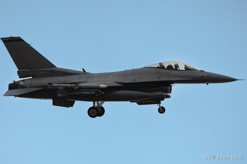 A gray F-16 fighter jet with landing gear down, underwing missiles and a fuel tank, flying left to right against a clear blue sky.