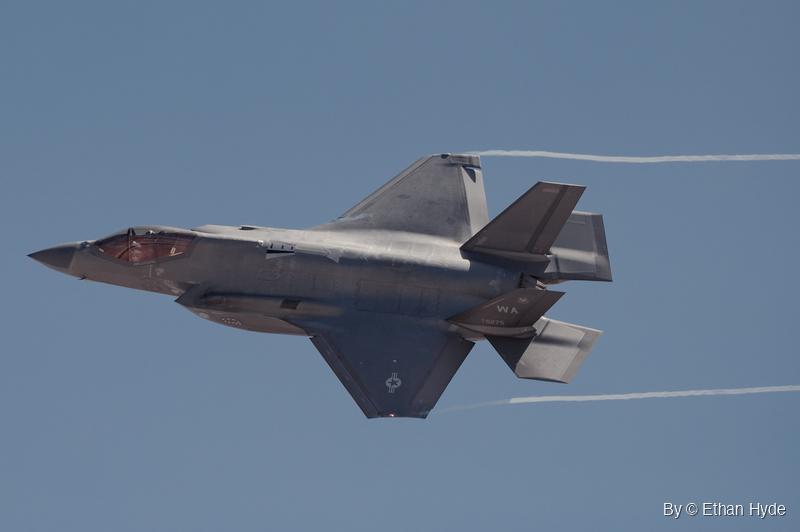 A gray F-35 fighter jet flies right to left in a banked turn with its underside showing, thin white vapor trails streaming behind it.