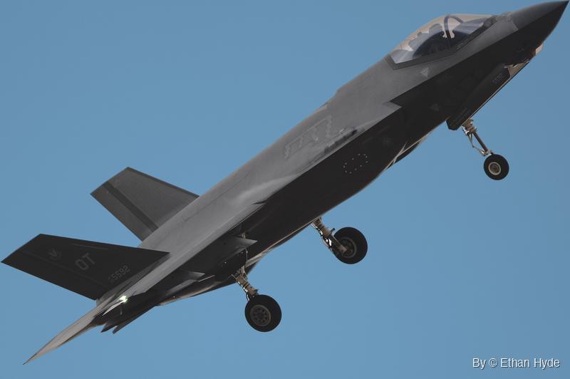 An F-35 fighter jet seen from below against a clear blue sky, landing gear extended and the pilot visible under the canopy.