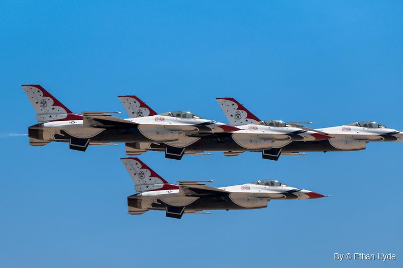 Four F-16 fighter jets in red, white and blue demonstration paint fly in tight formation, three overlapping in a line and one below, against blue sky.