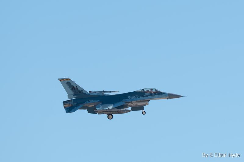 An F-16 in blue and gray pixelated camouflage flies with its landing gear down against a clear blue sky.