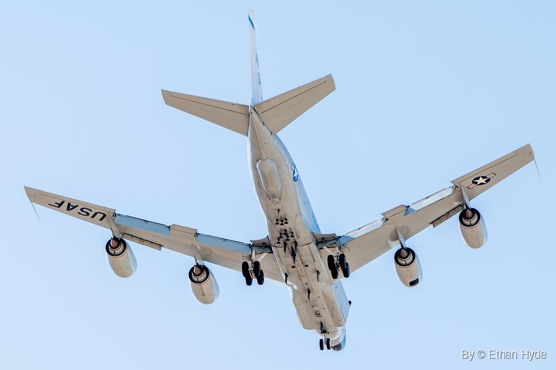 A white four-engine US Air Force RC-135 passes directly overhead with landing gear extended, USAF lettering and star insignia under the wings.