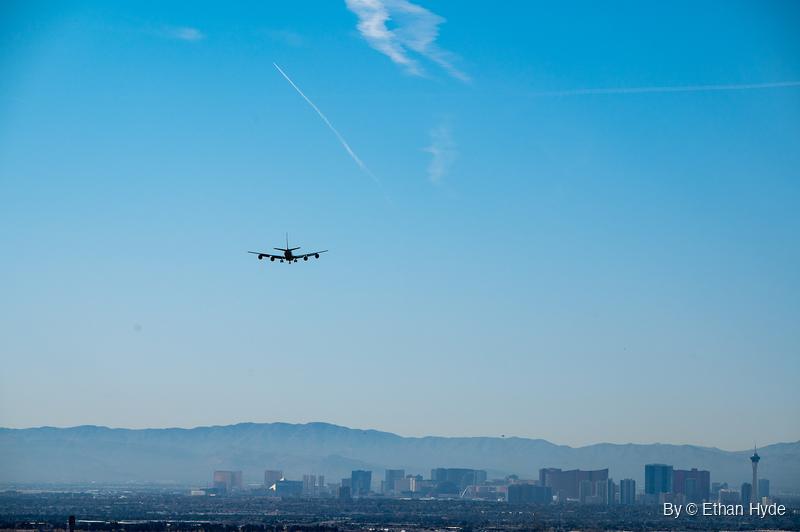 A four-engine RC-135 with landing gear down flies above a hazy skyline of high-rise towers, with a mountain range behind.